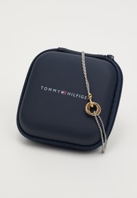 Custodia per gioielli Tommy Hilfiger blu navy con collana a catena argentata che presenta anelli intrecciati in oro su uno sfondo bianco.