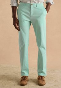 Világoszöld, egyenes szabású pamutnadrág, elöl zsebekkel és gombos záródással, barna fűzős cipőkkel párosítva.