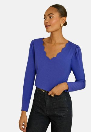 Blaues Langarmshirt mit einem gewellten Ausschnitt und gepufften Ärmeln, kombiniert mit dunklen Jeans aus Denim. Glatter Stoff mit einer figurbetonten Passform.