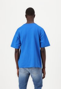 Μπλούζα από βαμβάκι σε μπλε χρώμα με κοντά μανίκια, χαλαρή εφαρμογή και λαιμόκοψη στρόγγυλη. Φοριέται με ανοιχτόχρωμα μπλε τζιν, εμφανιζόμενη από πίσω.