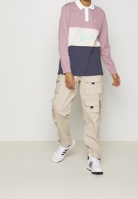 Langärmliges Polo in Pink, Weiß und Marineblau mit gesticktem Logo. Kombination mit lockeren beigen Cargo-Hosen mit Taschen und Reißverschlüssen. Schwarz-weiße Sneaker.