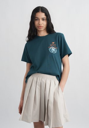 Kobieta z długimi ciemnymi włosami, ubrana w turkusowy T-shirt z grafiką NYC i beżową plisowaną spódnicę, stojąca z jedną ręką w kieszeni na jednolitym tle.