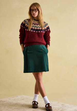 Burgundy knitted sweater with a geometric pattern, paired with a green corduroy skirt featuring double buttons and pockets. Brown shoes and white socks.
