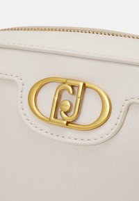 Borsa in pelle bianca con un accento in metallo dorato a forma circolare e un logo stilizzato, texture liscia e cuciture meticolose lungo i bordi.