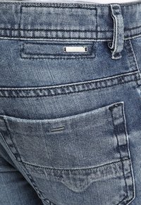 Blå denimjeans med en sliten textur, som har en stor bakficka och en metallisk logotypetikett. Exponerad sömdetalj längs sömmarna.