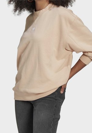 Osoba ubrana w beżową, oversize'ową bluzę z białym logo Adidas oraz ciemnoszare dżinsy, ręka spoczywa w tylnej kieszeni, gładkie tło.