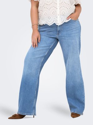 Kvinne iført lyseblå vidbente jeans, hvit bluse med hullbroderi, brune spisse hæler, med den ene hånden i lommen og den andre hvilende på låret.