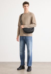 Zwarte crossbody tas, gladde textuur, gebogen vorm. Model draagt een beige trui en lichtblauwe jeans, staand op een neutrale achtergrond.