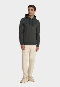 Felpa grigia scura con grafica frontale, coulisse regolabile, polsini e orlo a coste; abbinata a pantaloni chiari e sneakers beige.