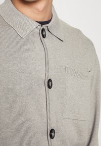 Lichtgrijze gebreide cardigan met zwarte knopen en een borstzak, gedragen over een zwart kledingstuk, cropped bij de nek en borst.