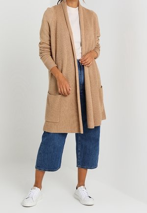 Femme portant un long cardigan beige avec des poches, un haut blanc, un jean court bleu et des baskets blanches, debout devant un fond blanc.