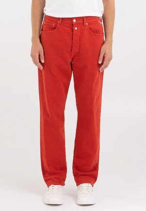 Pantalones de algodón rojos con corte recto, que cuentan con cinco bolsillos, herrajes en tono dorado y costuras en contraste. Llevados con zapatillas blancas.