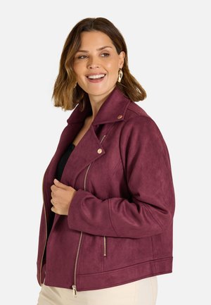 Frau lächelt zur Seite, trägt goldene Creolen, eine dunkelviolette Jacke mit Reißverschlüssen und hellfarbene Hose vor neutralem Hintergrund.