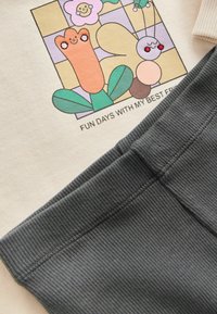 Gräddfärgad skjorta med en tecknad grafik och text, i kombination med mörkgrå ruggade byxor med en slät midja och textur.