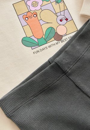 Camicia color crema con una grafica di cartone animato e testo, abbinata a pantaloni a coste grigio scuro con una cintura liscia e una texture morbida.