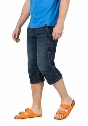Dunkelblaue Denim-Capri-Shorts mit mehreren Taschen und einem Kordelzug-Detail. Kombiniert mit orangen Sandalen mit offenem Zeh und einem blauen T-Shirt.