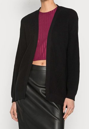 Woman wearing a black open-front knit cardigan over a purple cropped top and black leather skirt, standing against a plain background.