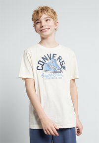 T-shirt en coton couleur crème avec un graphique bleu "Converse Original All Star" incluant un design de basket et une date d'établissement, à manches courtes.
