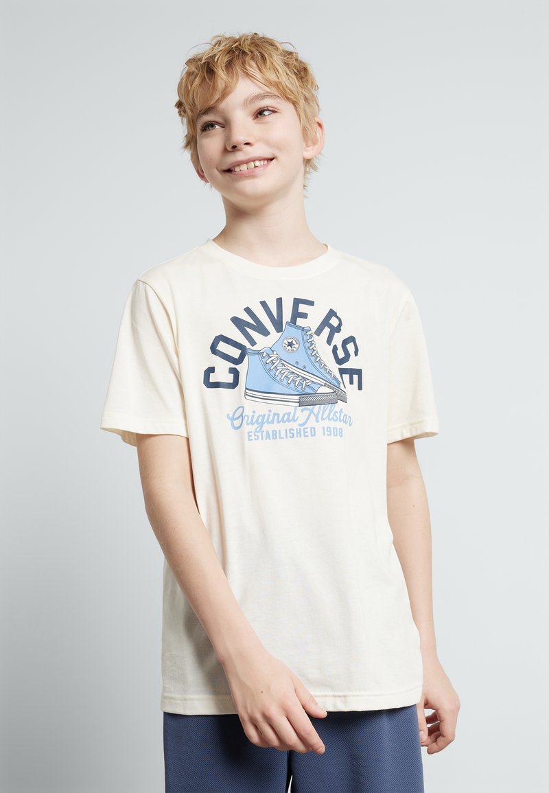 T-shirt en coton couleur crème avec un graphique bleu "Converse Original All Star" incluant un design de basket et une date d'établissement, à manches courtes.