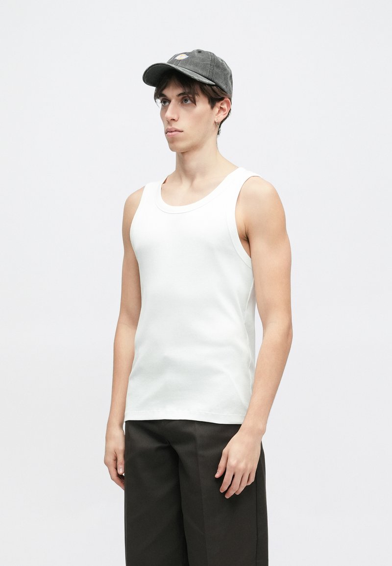 Hvid ribbet tanktop med rund hals, parret med mørke bukser. Modellen bærer en grå kasket med et broderet logo.