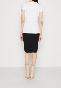 Mujer vista desde atrás, que lleva una blusa blanca de manga corta, una falda lápiz negra hasta las rodillas con abertura en la parte trasera y botines de tacón beis.