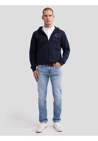 Férfi sötétkék cipzáras kapucnis pulóverben, fehér galléros pólóban, világoskék farmerben, fehér sportcipőben, egyszínű szürke háttér előtt áll.