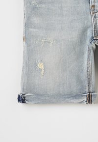 Jeans in denim azzurro chiaro con dettagli strappati, sfilacciature visibili e orli arrotolati. La texture è liscia; il tessuto appare ben consumato.