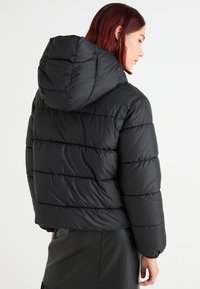 Veste rembourrée noire avec capuche, dotée de rayures matelassées horizontales et de poignets élastiques. Le matériau semble brillant et lisse.