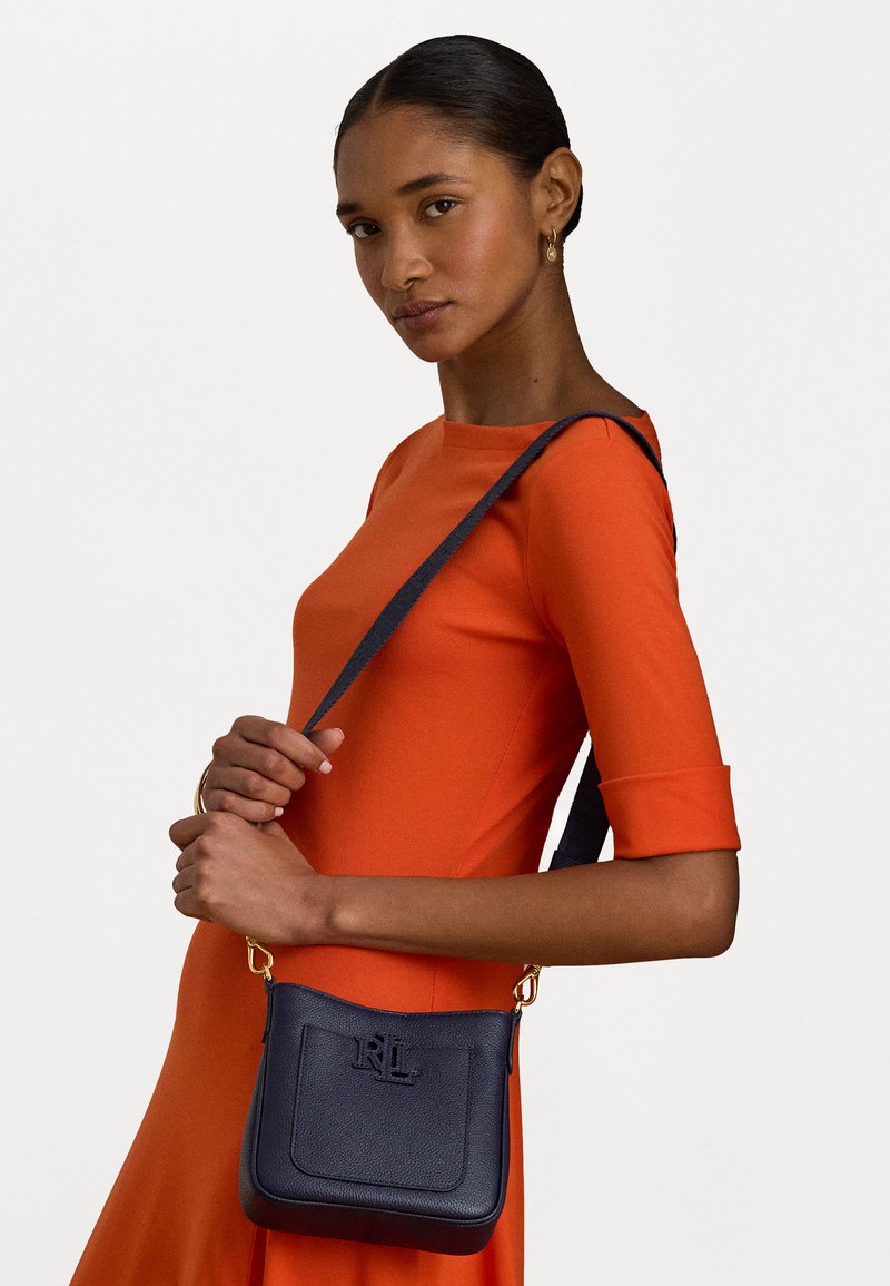 Tumši zilas krāsas ādas crossbody soma ar teksturētu virsmu, zelta aksesuāriem, iegravētu logotipu un regulējamu siksnu. Ietver priekšējo kabatu.