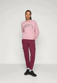 Donna in piedi con le mani dietro la schiena, indossando un felpa rosa DKNY, pantaloni cargo bordeaux, calze bianche e sneakers nere su uno sfondo neutro.