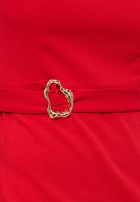 Κόκκινη υφασμάτινη ζώνη με ακανόνιστη, λαμπερή χρυσή αγκράφα, σφίγγοντας το ύφασμα στη μέση.