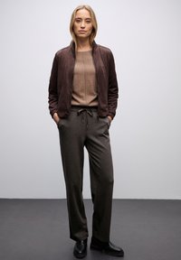Hnědá semišová bunda s postaveným límcem, nošená přes béžový svetr. Pří look dotváří tmavě šedé kalhoty se stahovací šňůrkou a černé kotníkové boty.