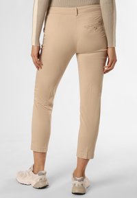 Kobieta w beżowych, dopasowanych do kostek spodniach z tylną kieszenią zapinaną na guzik, beżowym swetrze o prążkowanej strukturze i beżowych sportowych butach na grubej podeszwie, stojąca na białym tle.