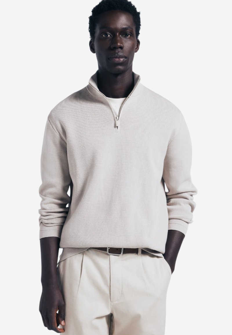 Maglione beige chiaro con colletto a zip quarti, polsini a coste e finitura strutturata, abbinato a pantaloni crema e una cintura grigia.