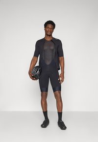 Schwarzes Radsporttrikot mit kurzen Ärmeln über einem schwarzen Mesh-Unterhemden, schwarze gepolsterte Shorts und einen schwarzen Helm, der in einer Hand gehalten wird.