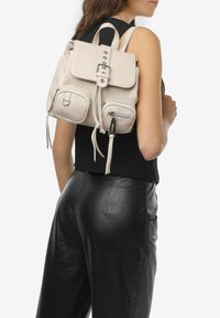 Beżowy skórzany plecak z klapą, dwoma przednimi kieszeniami na zamek i srebrnymi akcentami, noszony na plecach. Gładka tekstura i proste linie.