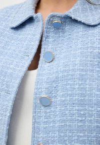 Giacca texturizzata blu chiaro con colletto e cinque bottoni tondi con centri blu e bordi dorati, creando un design strutturato e informale.