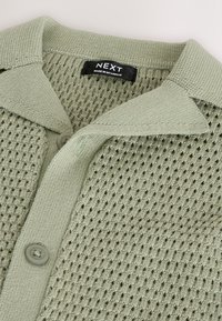 Lichtgroen gebreid vest met een gestructureerd patroon, knoopsluiting en een kraag. Label met de tekst "NEXT, gemaakt in Myanmar."