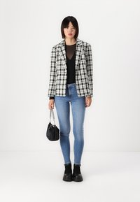 Juodas ir baltas houndstooth rašto bleizeris virš permatomos juodos bluzos, derinamas su šviesiai mėlynomis skinny džinsomis ir juodais auliniais batais, laikant juodą rankinę.