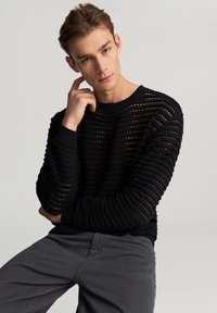 Giovane uomo con capelli castano chiaro, che indossa un maglione nero a maglia aperta e pantaloni grigi, seduto con una mano che tocca il collo.