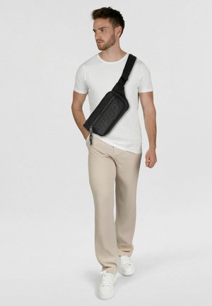 Hombre con camiseta blanca, pantalones beige acanalados, zapatillas blancas y bolso cruzado negro con el logo "HUGO", caminando frente a un fondo liso.