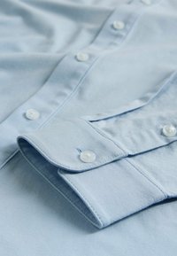 Gai zila krekla ar gludu tekstūru, ar vienu pogu aprocei un pērļu līdzīgām pogām gar priekšpusi.