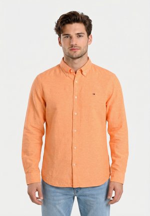 Uomo che indossa una camicia arancione a maniche lunghe con bottoni e jeans azzurri, in piedi davanti a uno sfondo bianco uniforme.