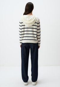 Gestreifter Kapuzenpullover in Creme mit marineblauen Streifen, kombiniert mit lockeren blauen Jeans und weißen Turnschuhen, von hinten betrachtet.