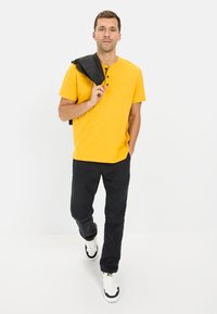 Gul kortærmet henley-skjorte med tre knapper, parret med sorte bukser og hvide sneakers, iført en sort jakke over skulderen.