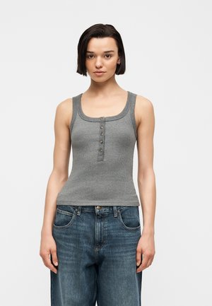 Femme aux cheveux courts et foncés portant un débardeur gris côtelé sans manches avec des boutons et un jean bleu ample, debout devant un fond uni.