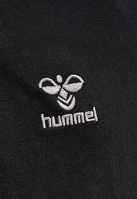Tessuto nero con un logo ricamato bianco di un'ape sopra il testo "hummel", con una texture liscia e dettagli chiari.