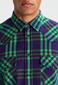 Fioletowa i zielona koszula w kratę z guzikami, posiadająca dwie przednie kieszenie, kontrastowe przeszycia oraz zapięcia na napy. Wyposażona w kołnierzyk w szpic i krótkie rękawy.