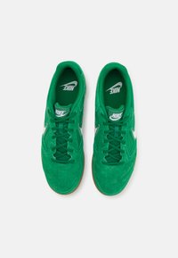Zeleni nubuk športni copati z belim Nike logotipom, z vezalkami, podloženim ovratnikom in podplatom iz gume. Spodnji pogled prikazuje podrobno teksturo.