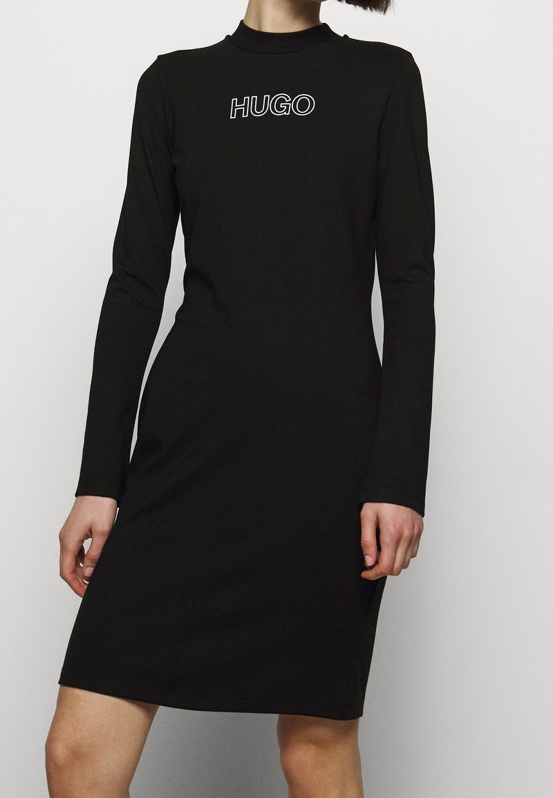 Femme portant une robe noire à manches longues, longueur genou, avec le logo "HUGO" imprimé en blanc sur la poitrine.